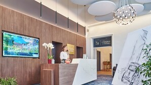 A reception area with a wooden reception desk, a TV screen displaying a scenic view, a reception desk with a sign, and a painting on easels.