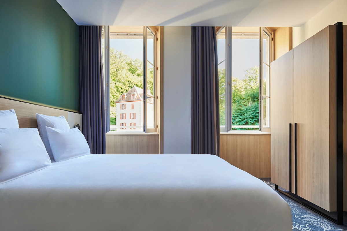 Hotellihuone, jossa on suuri sänky, vaatekaappi ja ikkuna, josta avautuu näkymä rakennukseen ja puihin.