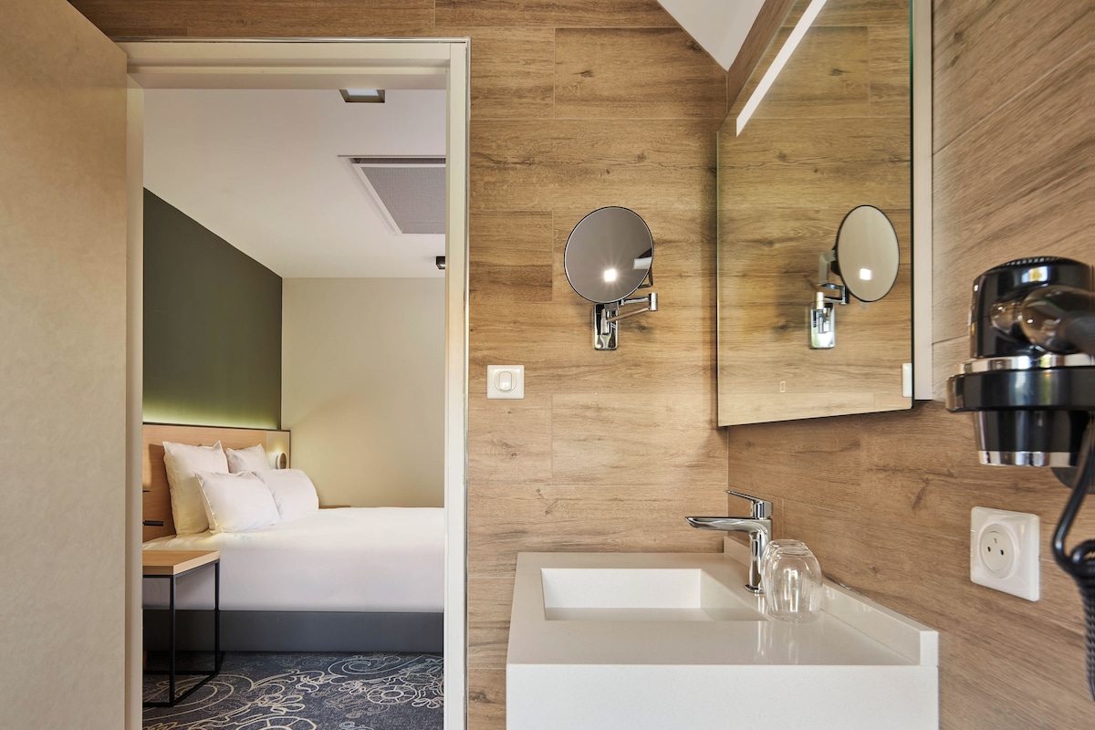 A modern hotel room with a wooden accent wall, a large mirror, a white sink, and a neatly made bed.