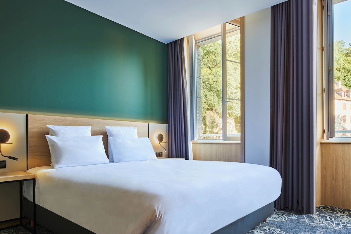 A hotel room with a large bed, a nightstand, a wall-mounted lamp, and a window with curtains.