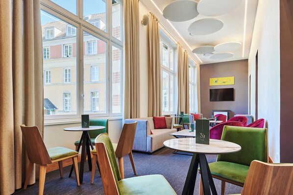 Una habitación de hotel moderna con ventanas amplias, un sofá, sillones, una mesita y un televisor de pantalla plana.