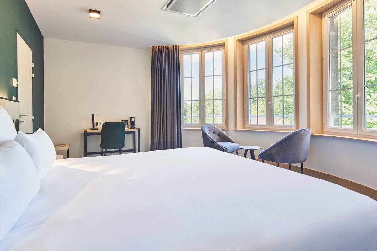 Una habitación de hotel con una cama grande, dos sillones, un escritorio y una ventana con cortinas.