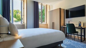 A hotel room with a bed, a desk with a chair, a TV, and large windows.