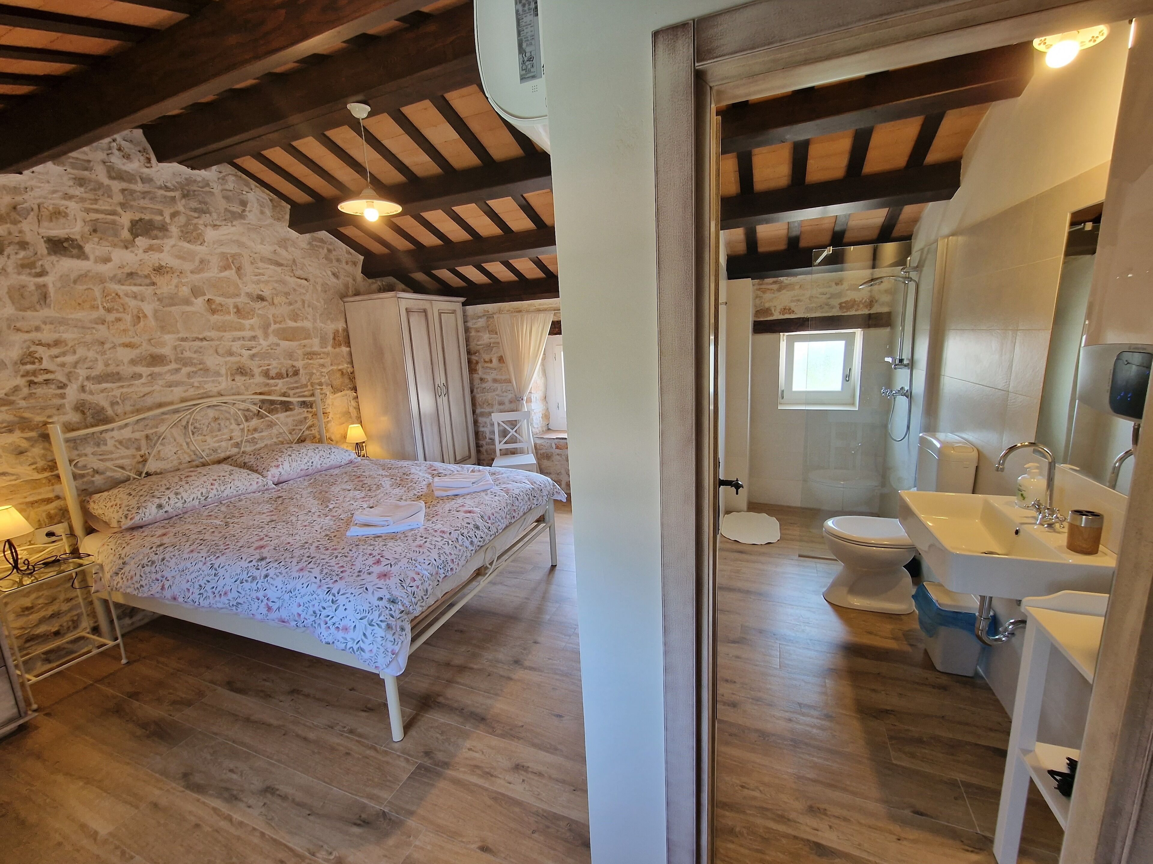 Une chambre avec un mur en pierre, des poutres en bois au plafond, un lit avec une literie à motifs floraux, une table de chevet et une commode.