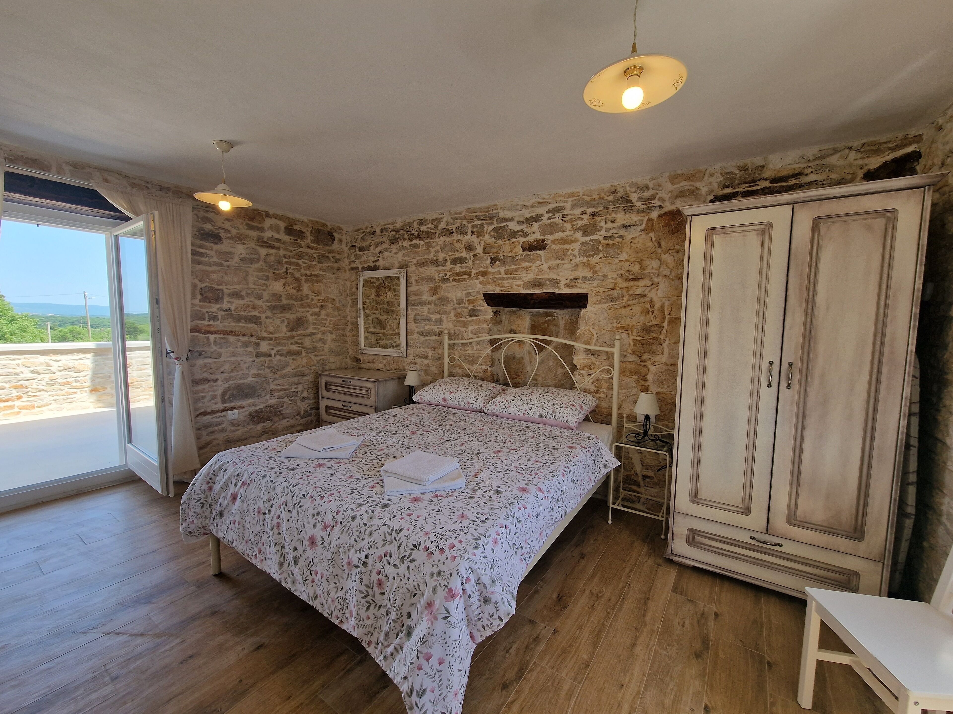 Une chambre avec un mur en pierre, un lit recouvert d’une literie à motifs floraux, une armoire en bois et une petite table.