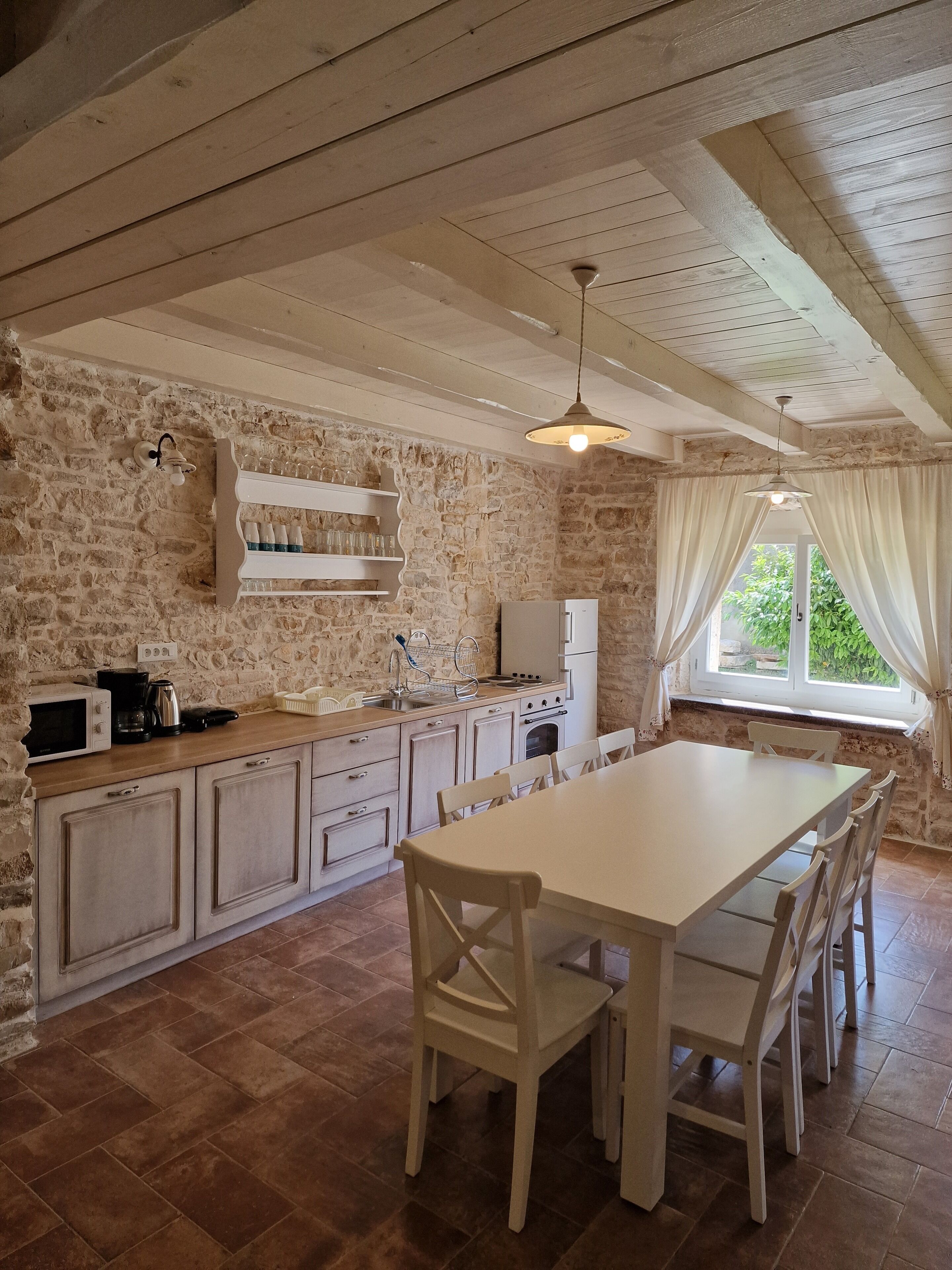 A rustic kitchen with exposed stone walls, wooden beams, and a large dining table with chairs.