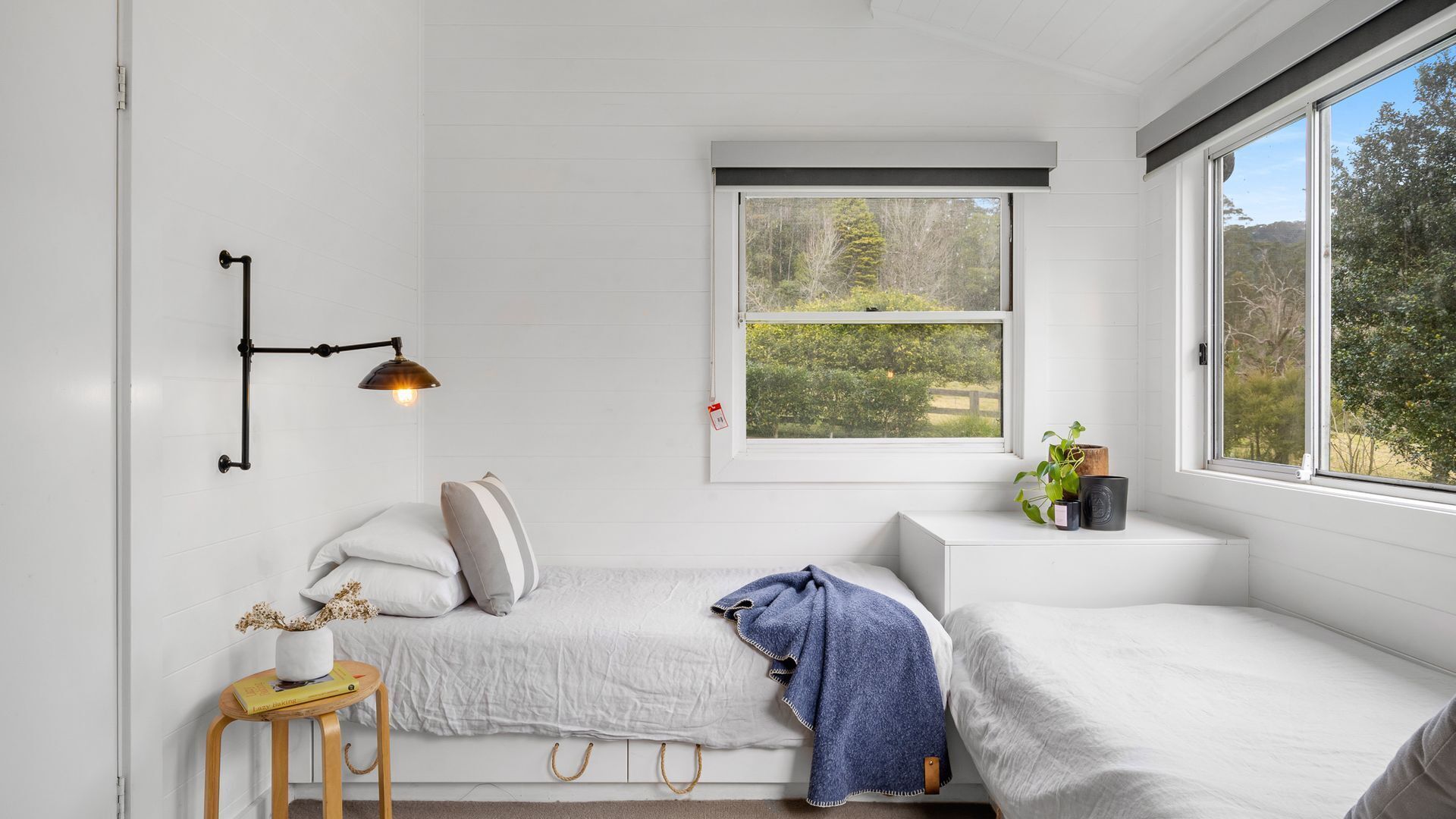 Et soveværelse med en seng, et sengebord med en lampe, et vindue med udsigt til grønne områder, og en lille reol med planter.