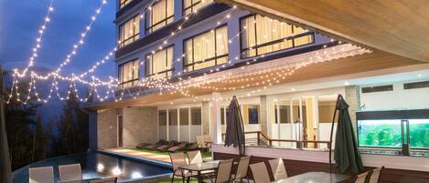 Un área de piscina al aire libre con sillas y mesas de descanso, luces colgantes y una zona de bar con un gran acuario.