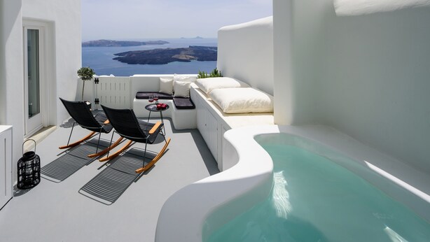 Une piscine à débordement blanche, de forme incurvée, deux chaises longues noires, une petite table sur laquelle se trouve un objet rouge, et une vue sur la mer et le paysage lointain.