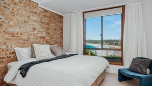 A bedroom with a brick wall, a large window with a view of the beach, a bed with striped bedding, and a blue chair. - Vista del Mare - Indulge yourself in the lap of luxury with endless ocean views (Mollymook Beach)