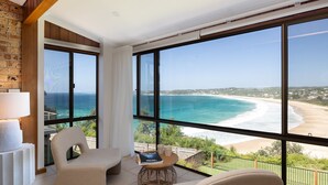 A room with large windows overlooking a beach, a chair, a table with a magazine, and a lamp. - Vista del Mare - Indulge yourself in the lap of luxury with endless ocean views (Mollymook Beach)