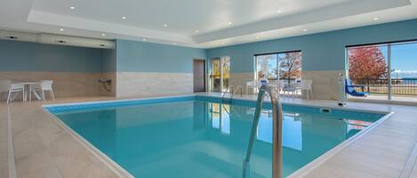 Una amplia piscina interior con agua cristalina y azul, rodeada de un suelo de baldosas y un área junto a la piscina con sillas.