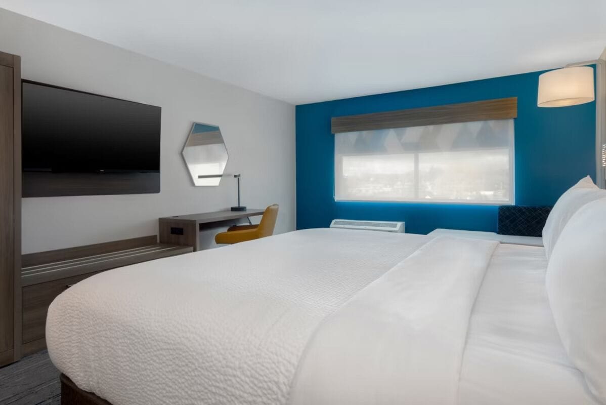 A hotel room with a large bed, a flat-screen TV mounted on the wall, a desk with a chair, and a window with a view.