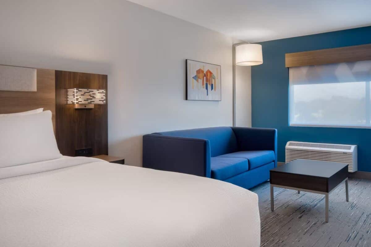 Ett hotellrum med en stor säng, en blå soffa, ett litet bord, en väggmonterad luftkonditioneringsanläggning och ett fönster med utsikt.