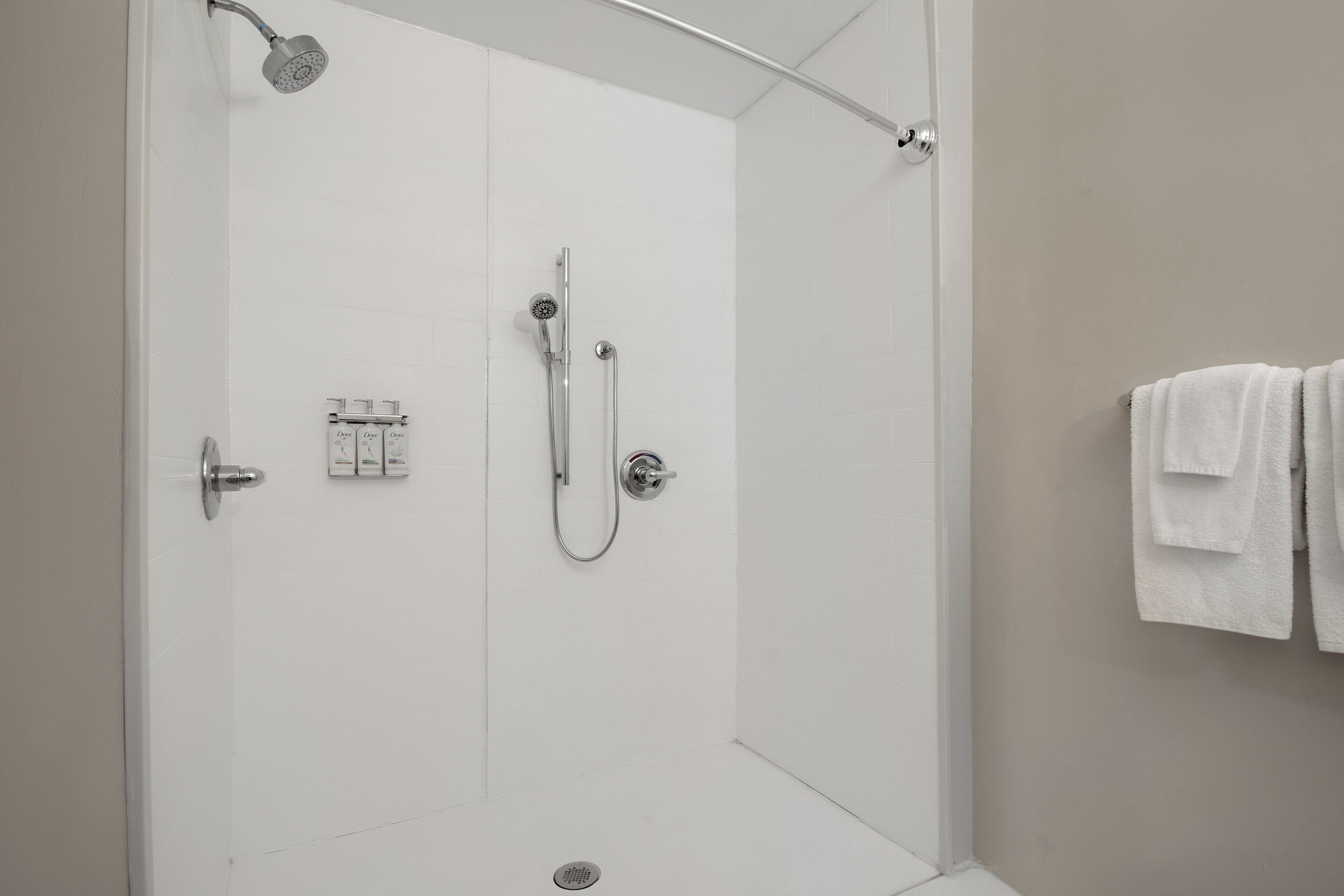 Ducha a nivel de suelo con mampara de vidrio, cabezal de ducha de mano, grifo empotrado en la pared y perchero con toallas.