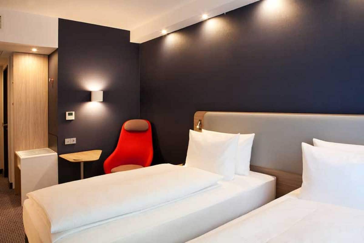 Une chambre d’hôtel avec deux lits, une chaise rouge, une petite table et un lampadaire fixé au mur.