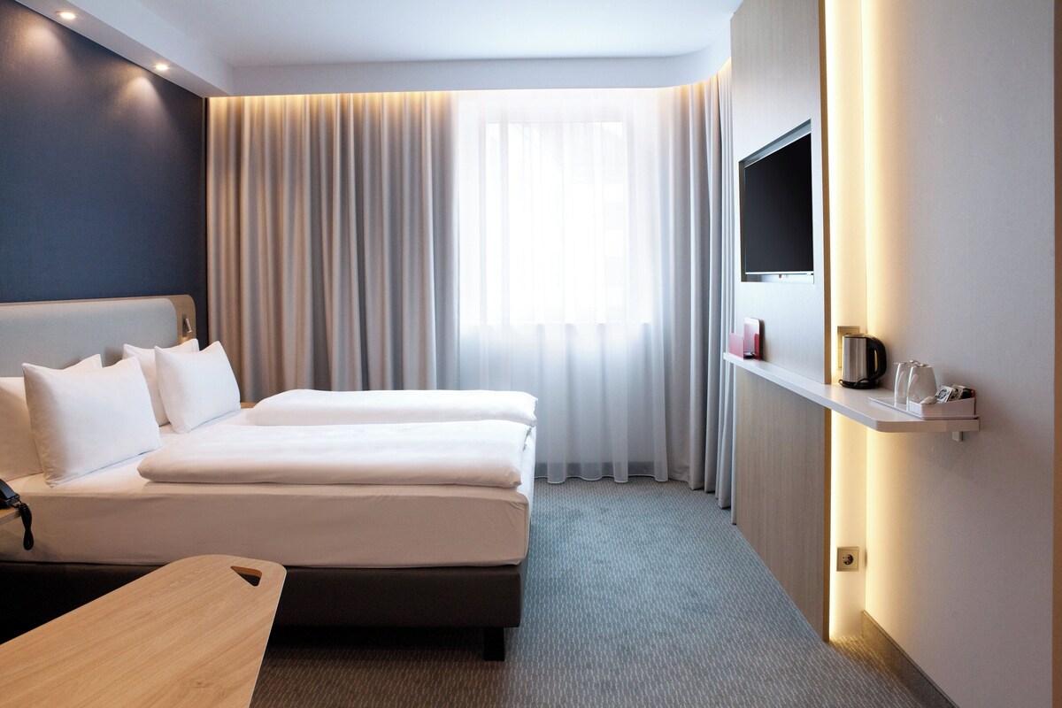 A hotel room with two beds, a desk, a TV, and a shelf with items.