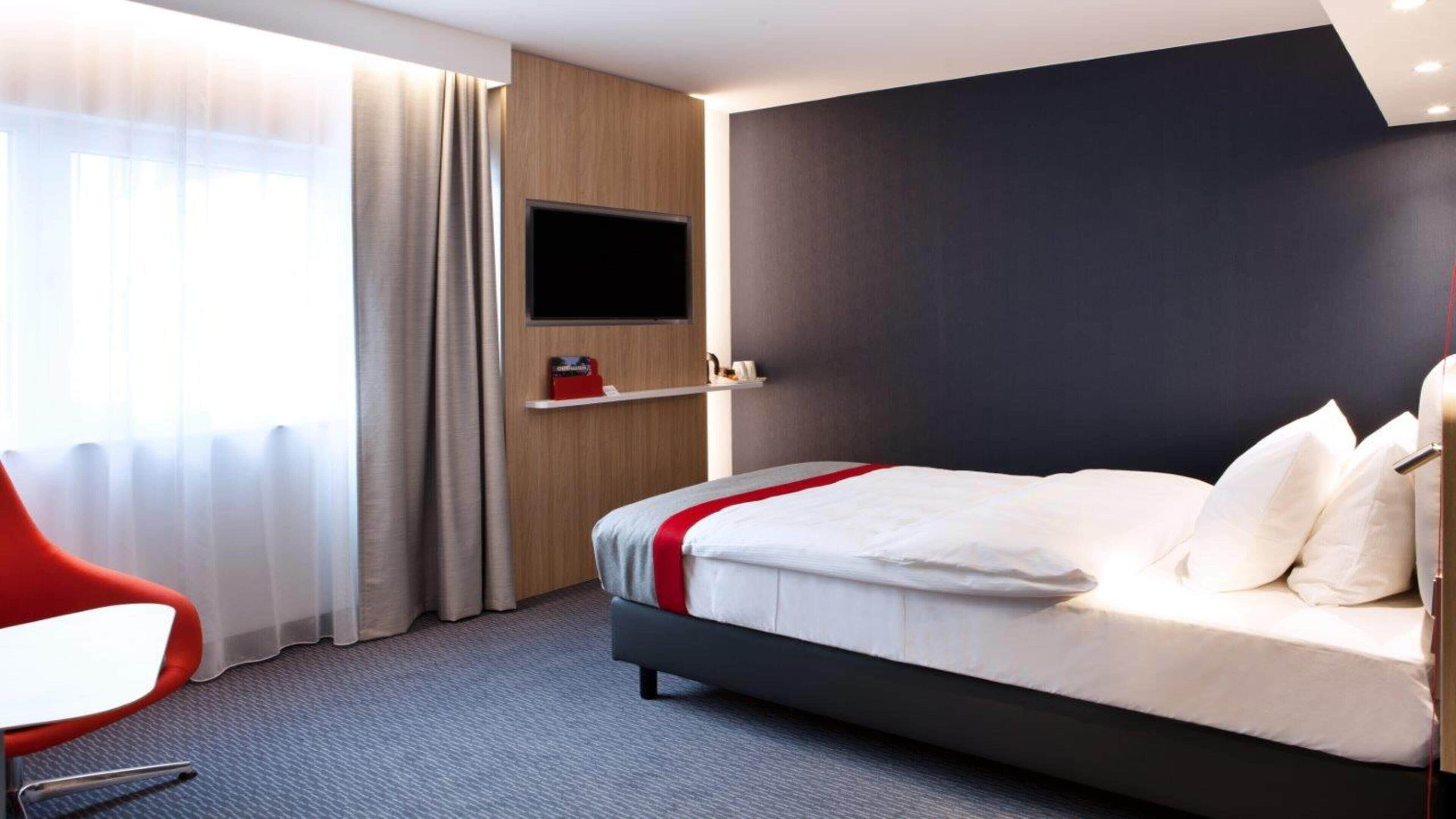 A hotel room with a bed, a TV mounted on the wall, a red chair, and a window with curtains.