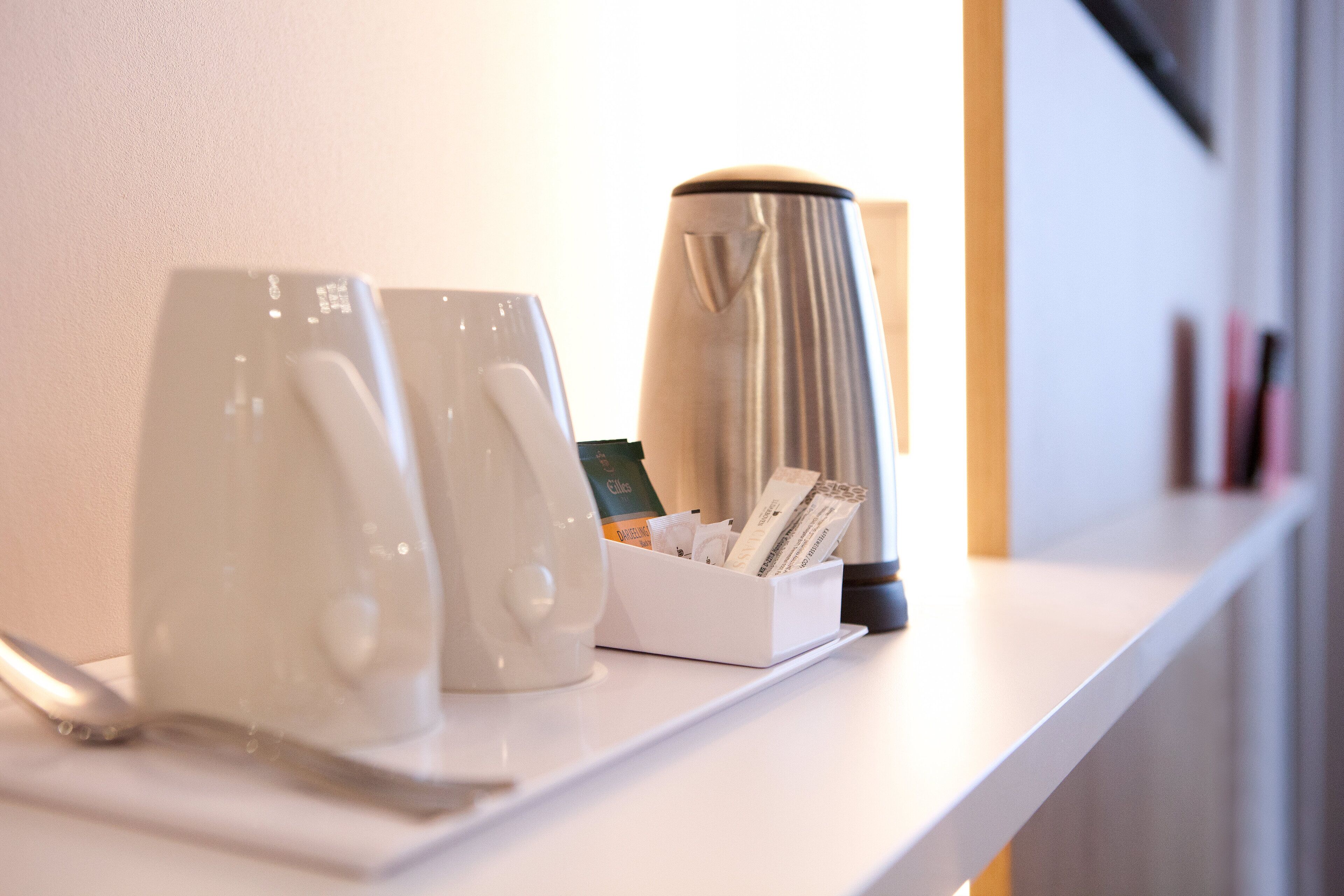 A white countertop with two white mugs, a white coffee pot, and a white box containing coffee cups and a spoon.