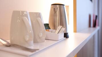 A white countertop with two white mugs, a white coffee pot, and a white box containing coffee cups and a spoon.