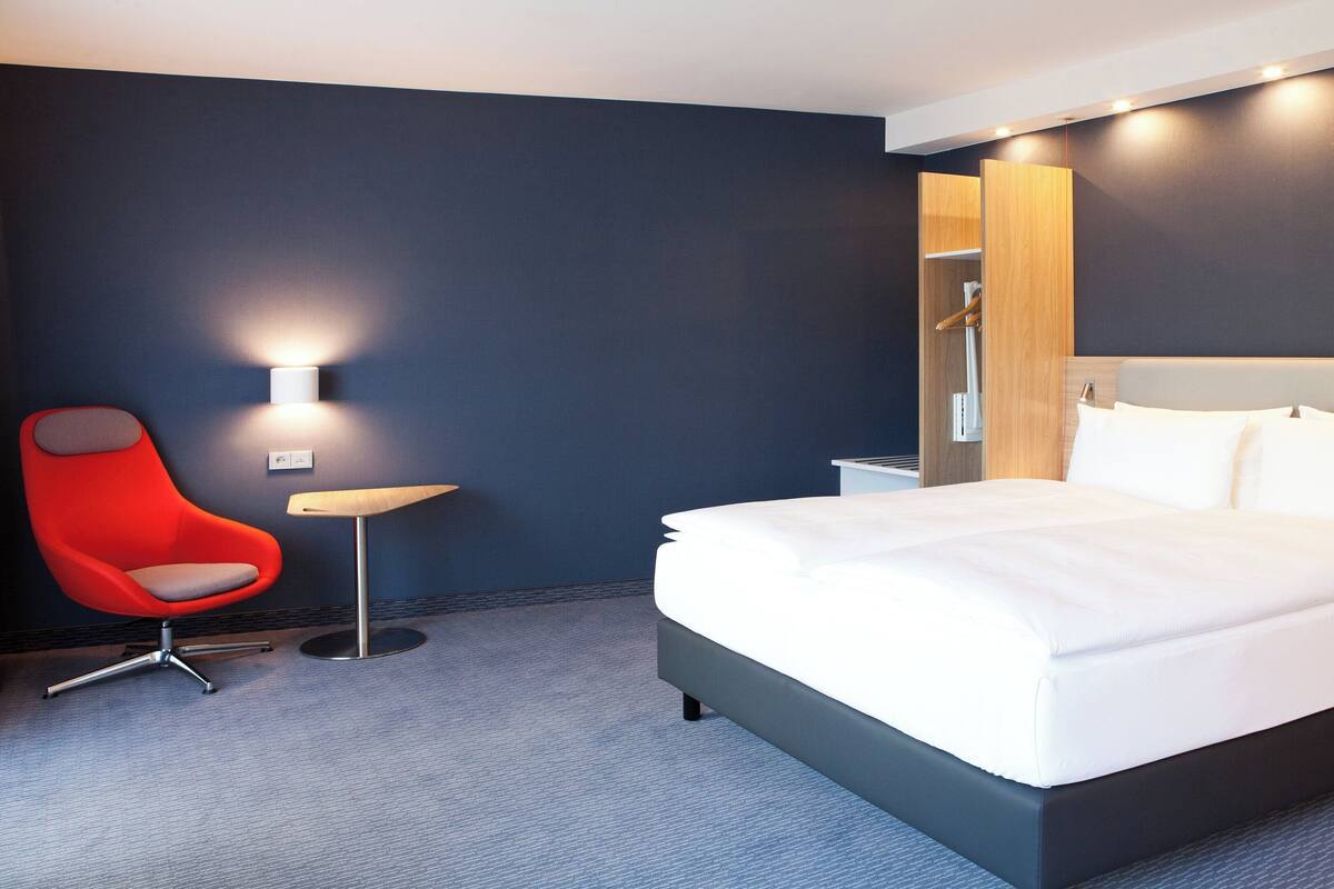 Une chambre d’hôtel moderne avec un grand lit, un fauteuil rouge, une petite table et un lampadaire fixé au mur.