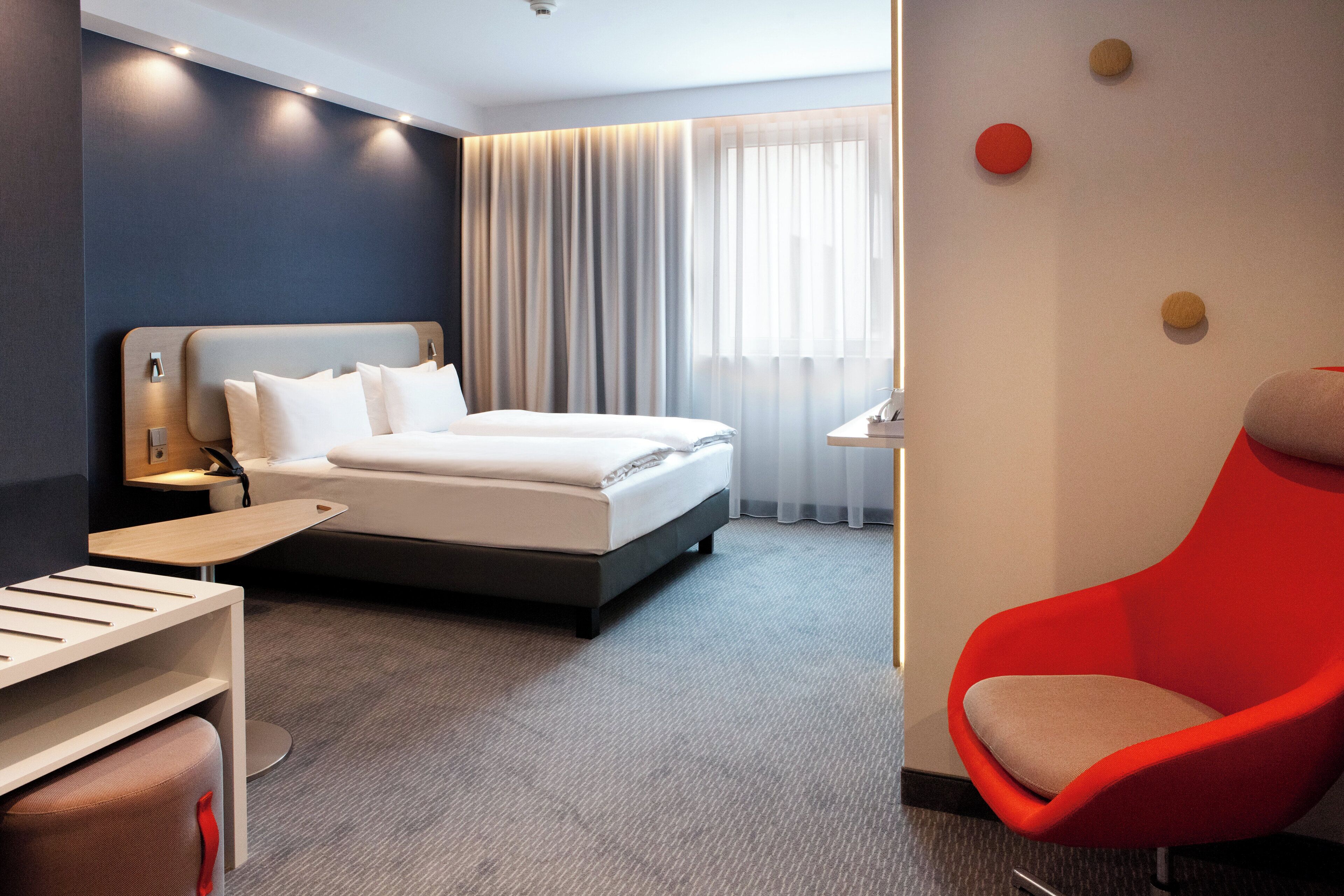 A modern hotel room with a bed, a desk, a red chair, and a large window with curtains.