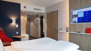 A modern hotel room with a large bed, a red chair, a small table, and a mounted TV displaying a room control interface. - Holiday Inn Express Bochum by IHG (Bochum)