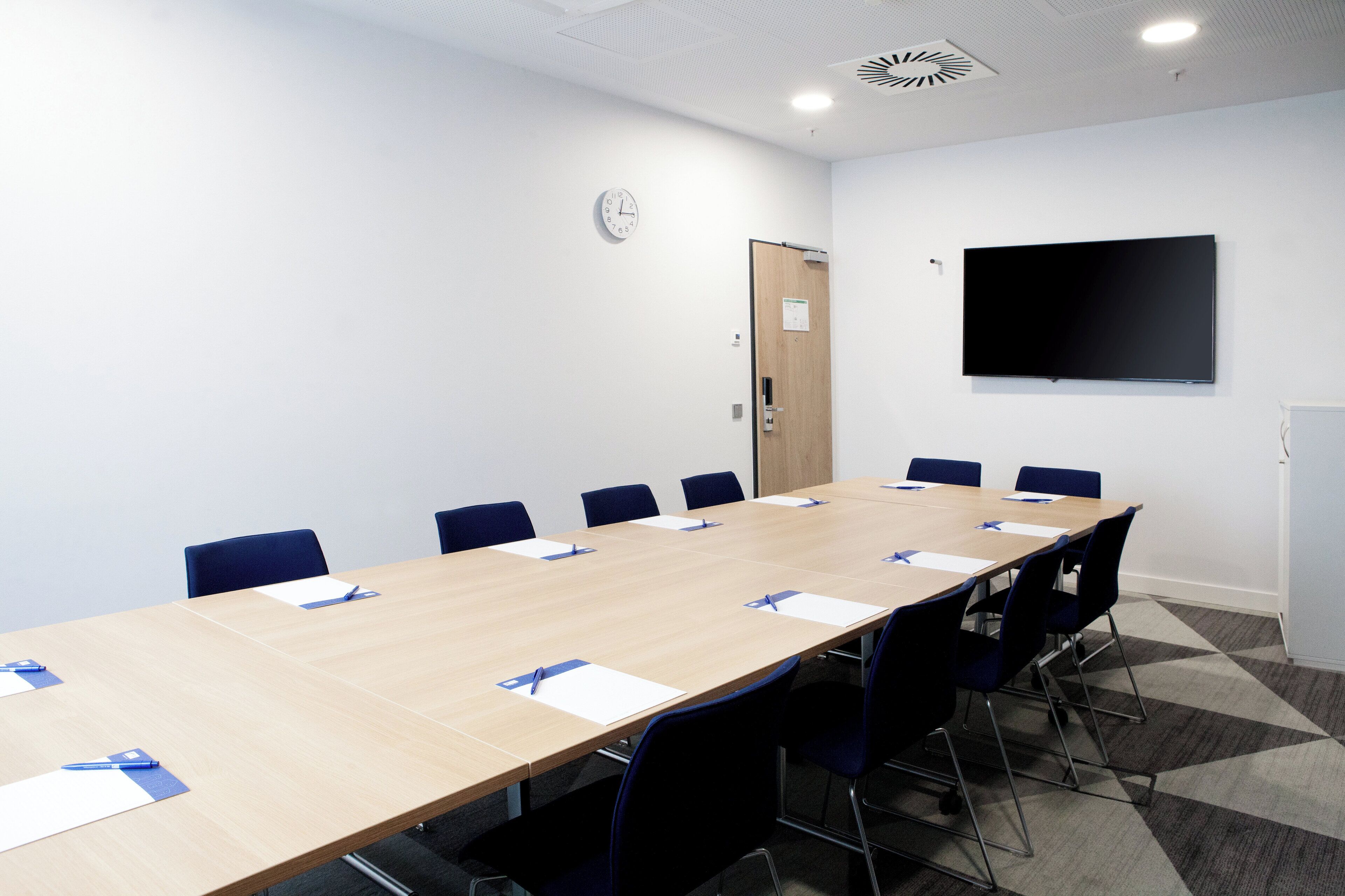A conference room with a long table, blue chairs, a clock, and a TV on the wall.