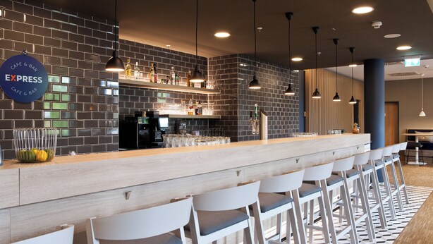 A modern bar area with a long counter, white bar stools, and a brick wall backdrop. - Holiday Inn Express Bochum by IHG (Bochum)
