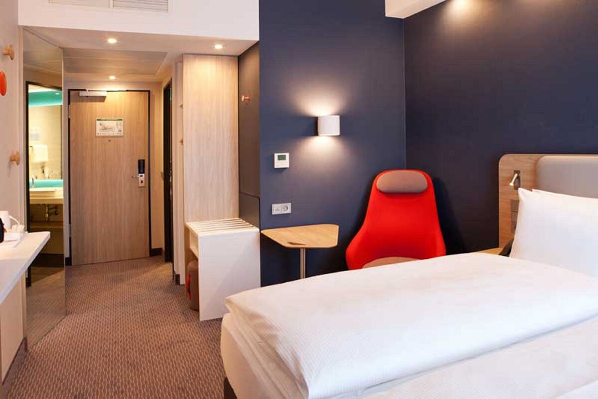 Una camera d'hotel con un letto, una sedia rossa, un tavolino, una scrivania e un bagno visibile tramite una porta aperta.