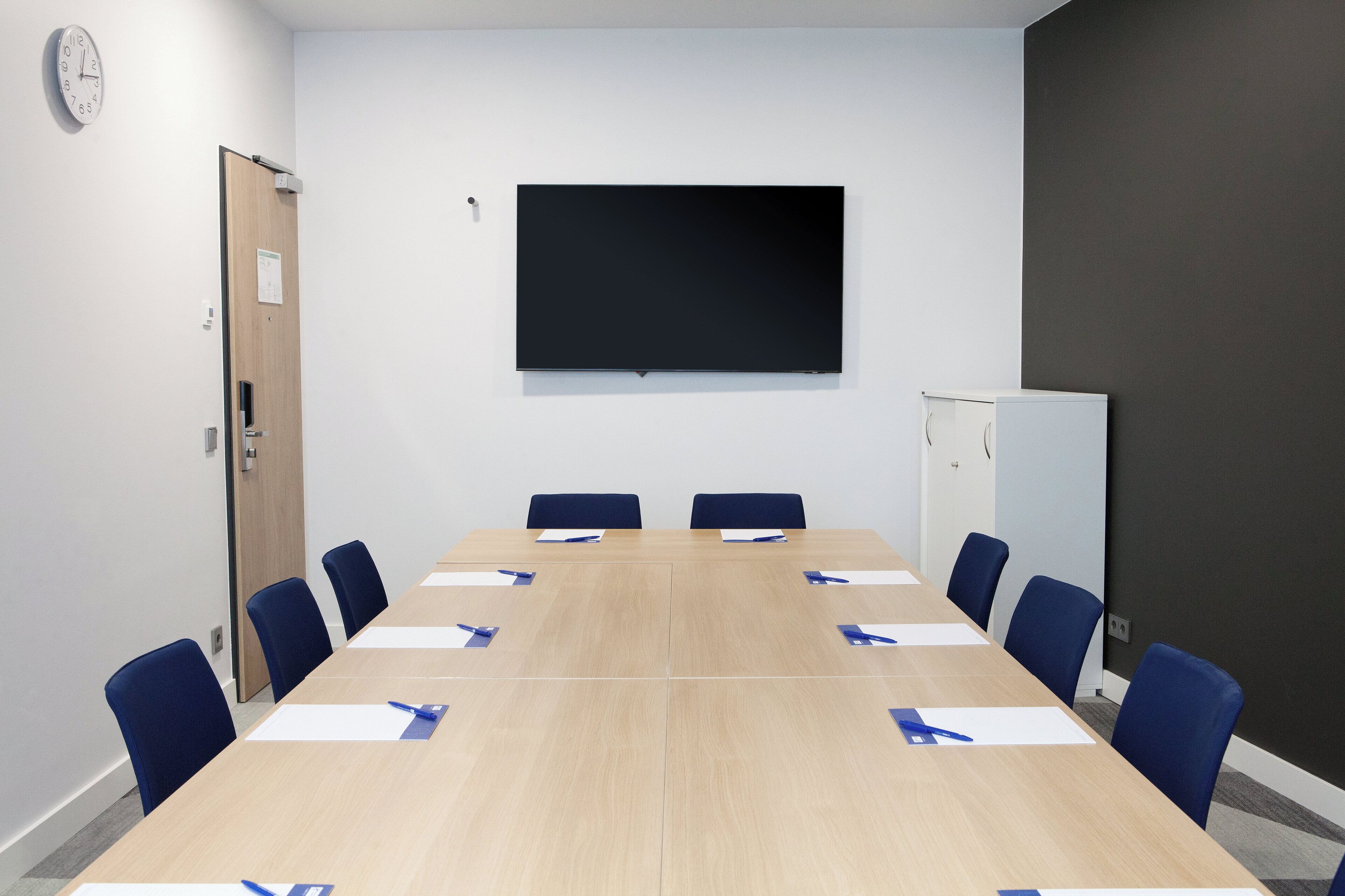Ein Konferenzraum mit langem Tisch, blauen Stühlen, einem Flachbildfernseher und einer Whiteboard.