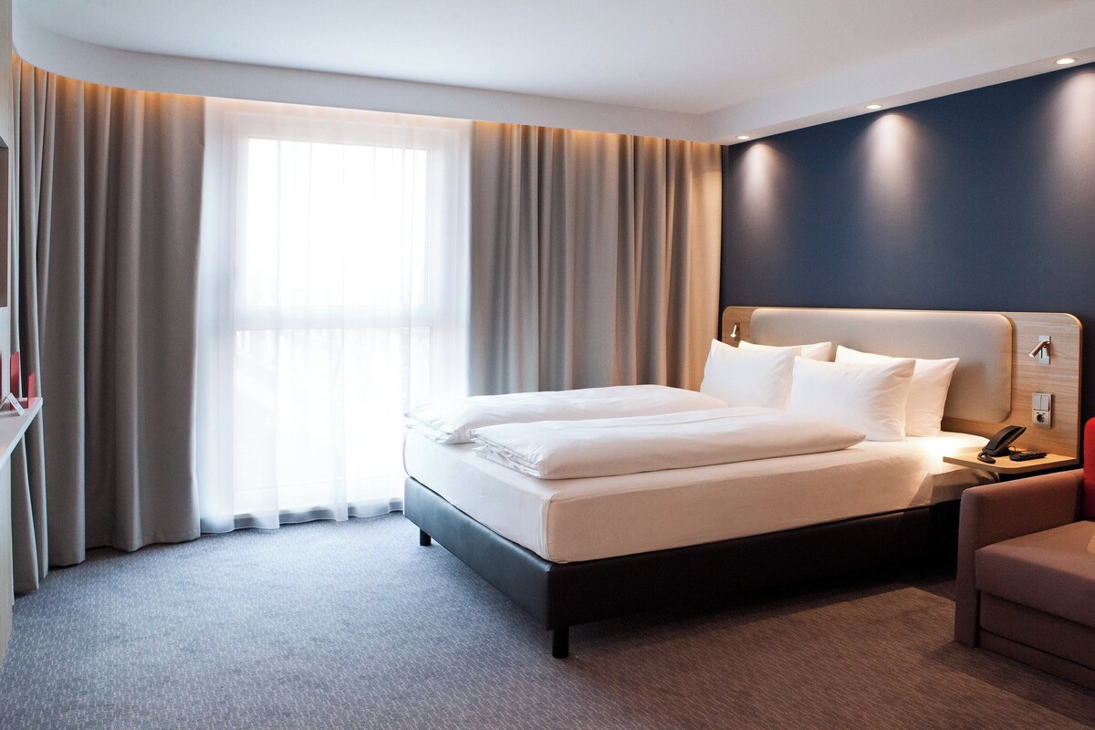 A modern hotel room with a large bed, a bedside table with a phone, a red chair, and a large window with curtains.