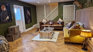 A living room with a brown leather sofa, a yellow armchair, a coffee table, and a lamp. There are two framed pictures on the wall, a staircase in the background, and a deer head mounted on the wall. - 8 Coach House (Preston)