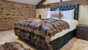 A bedroom with a stone wall, a tufted headboard, a fur throw, a bottle of champagne, and a suitcase. - 8 Coach House (Preston)