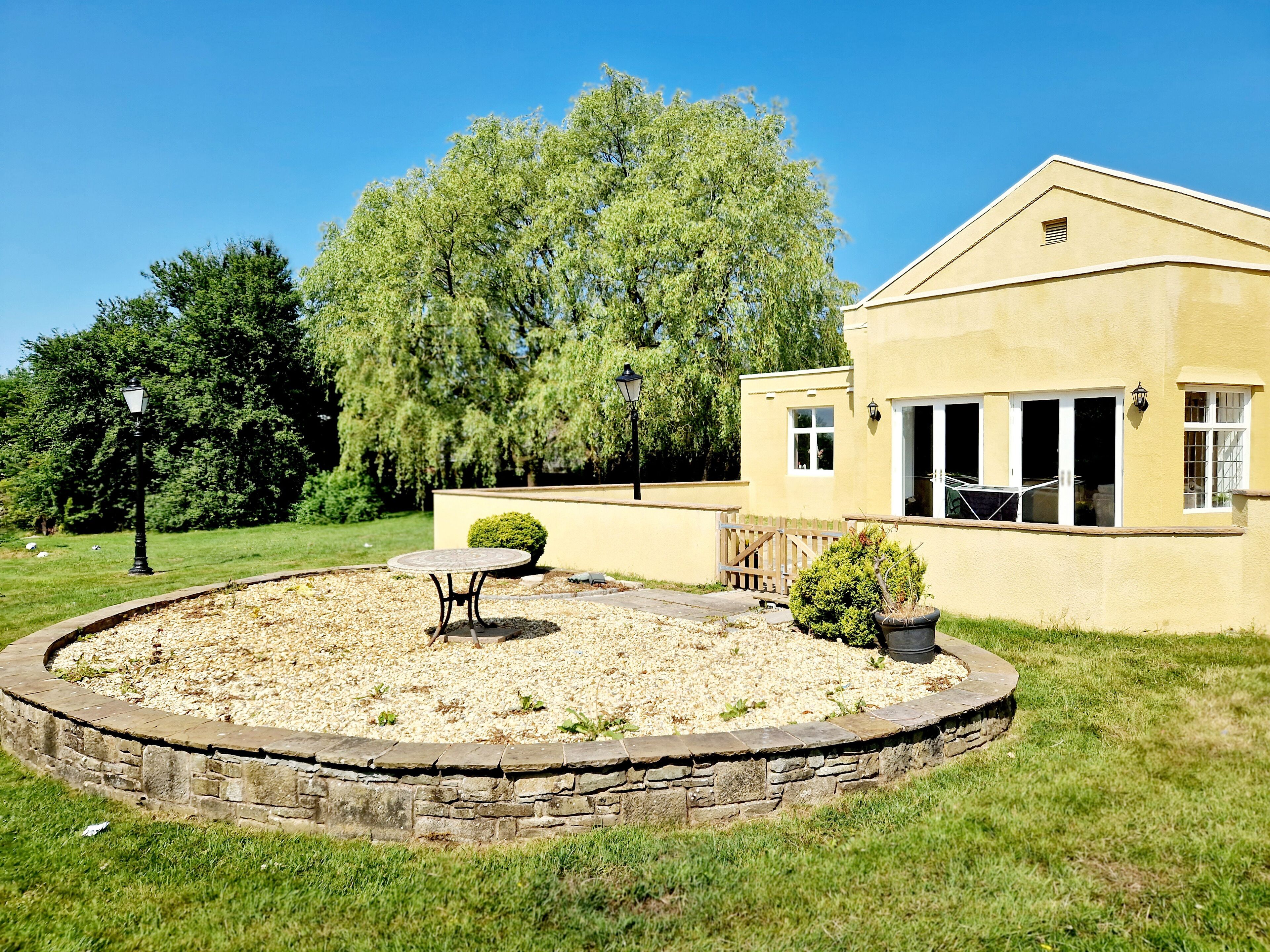 Une maison avec une terrasse circulaire en pierre, une table ronde et un petit arbuste.
