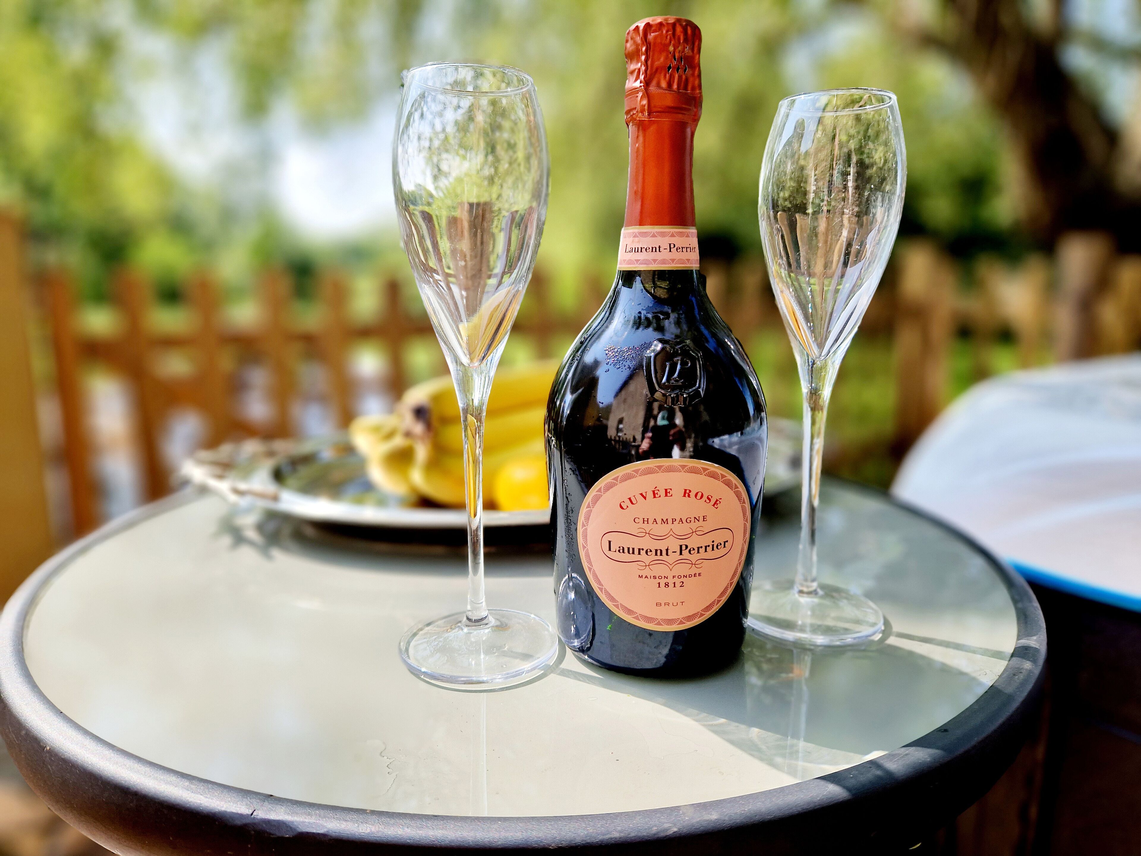 A bottle of Cuvée Rosé Champagne and two glasses on a round table with a blurred outdoor background.