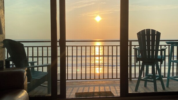 A room with a view of the sunset over the ocean, a sliding glass door, a brown leather sofa, and two green Adirondack chairs. - Stunning direct ocean front 2 bedroom unit! Fully equipped and all remodeled! (New Smyrna Beach)