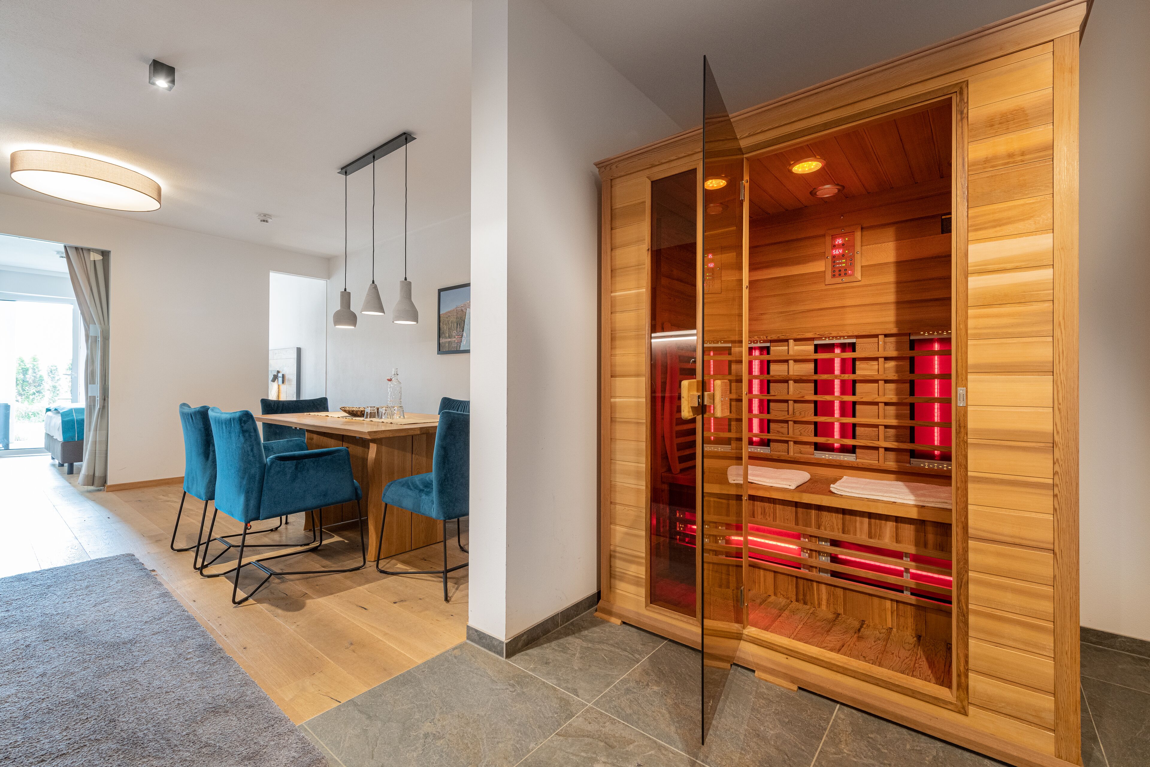 A modern hotel room with a wooden wardrobe, a dining area with blue chairs, and a large window.