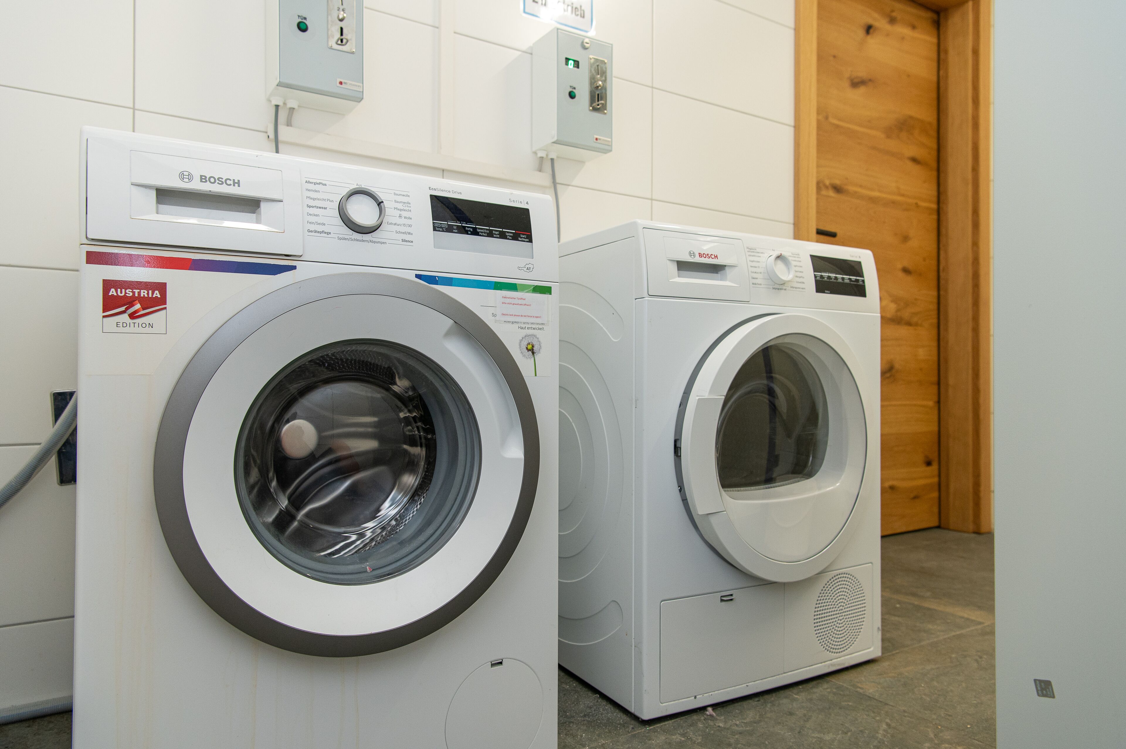 Two Bosch washing machines in a room with tiled walls and a wooden door.