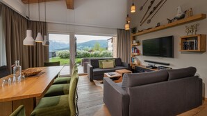 A modern living room with a wooden dining table, green upholstered chairs, a grey sofa, and a flat-screen TV mounted on the wall. - AlpenParks Hotel & Apartment Carpe Solem (Mariapfarr)