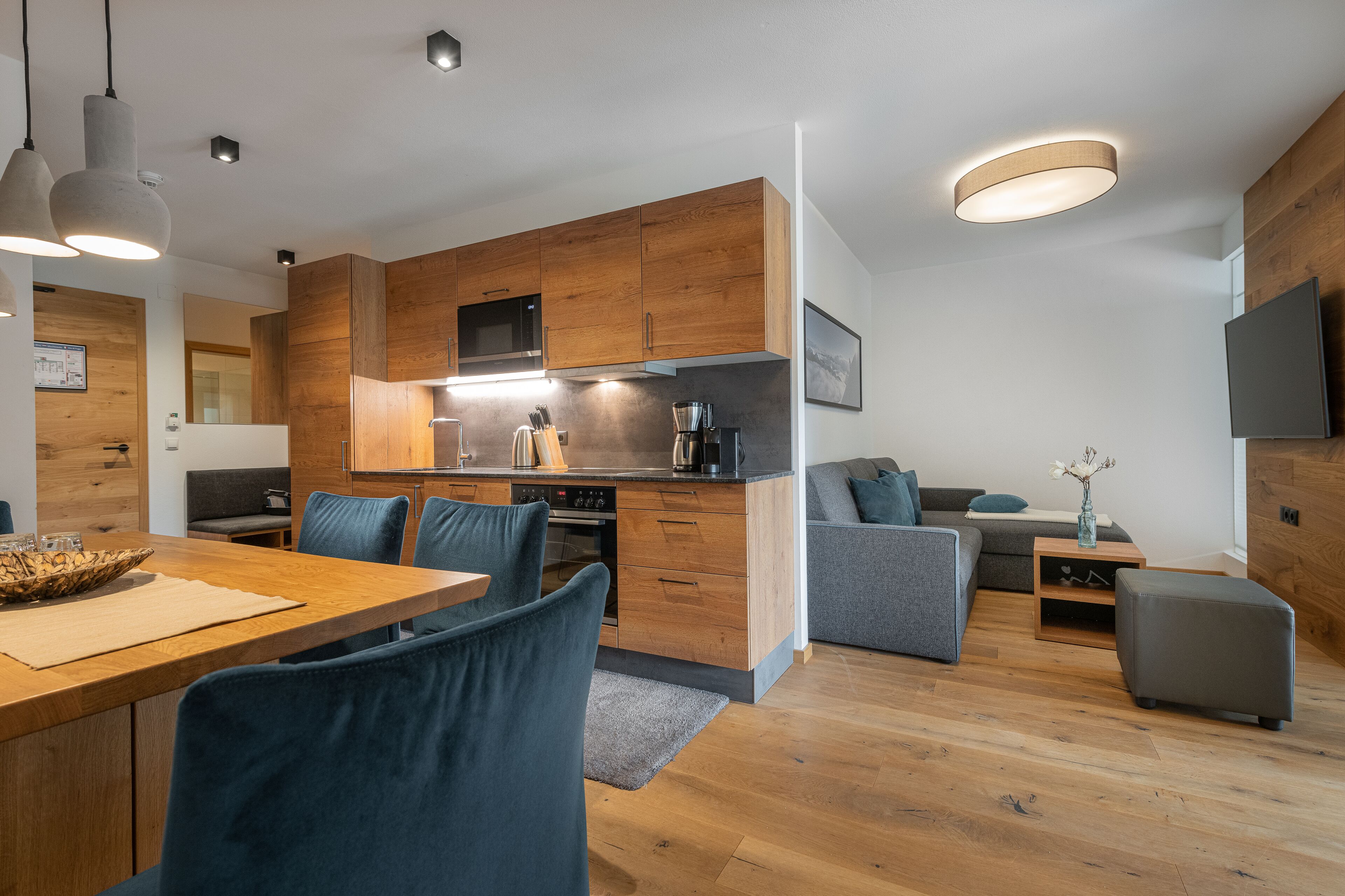 Eine moderne Küche mit Holzausstattung, ein Essbereich mit blauen Stühlen und ein graues Sofa in einem Wohnzimmer.