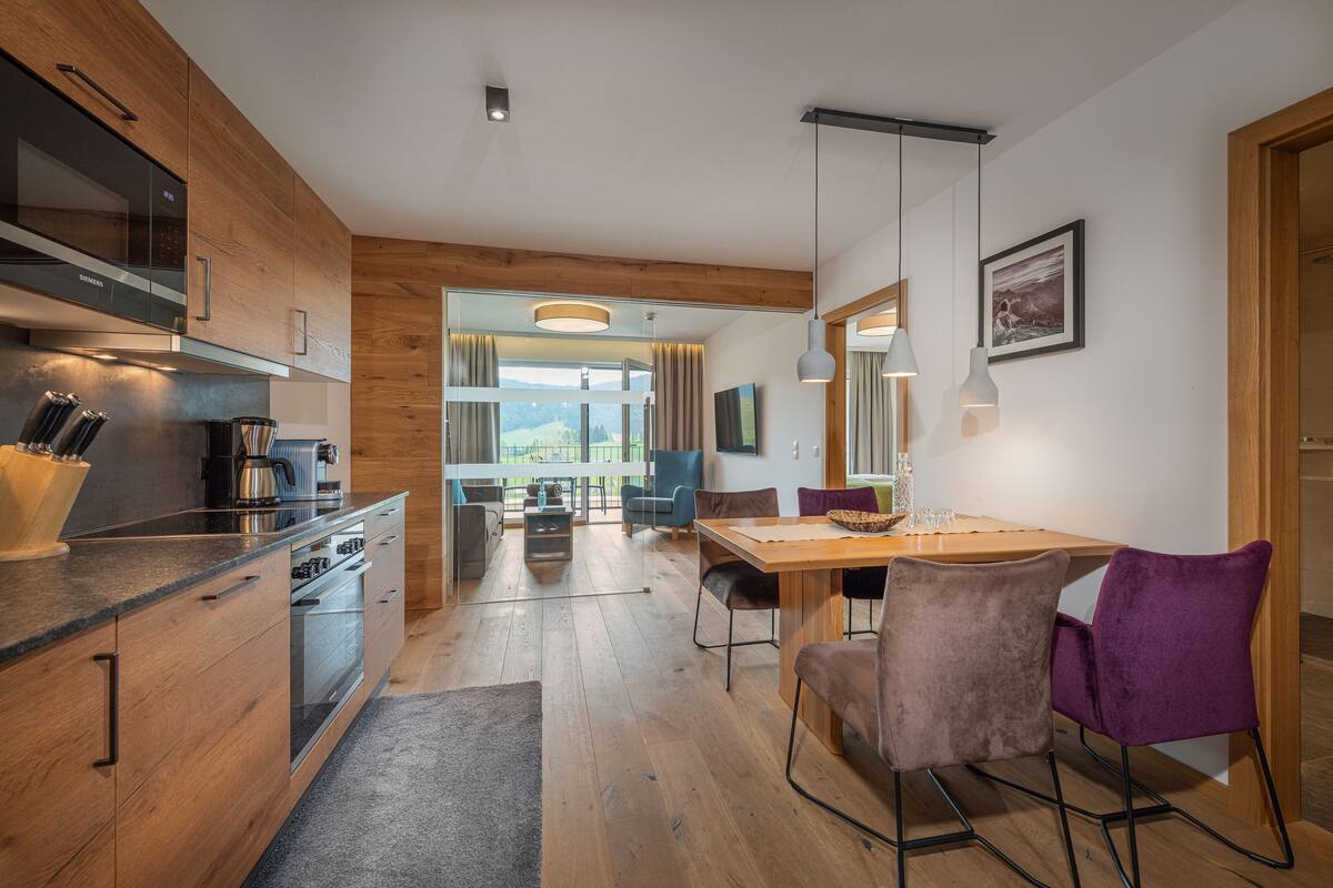 Cocina moderna con armarios de madera, zona de comedor con mesa y sillas, y vistas al exterior.