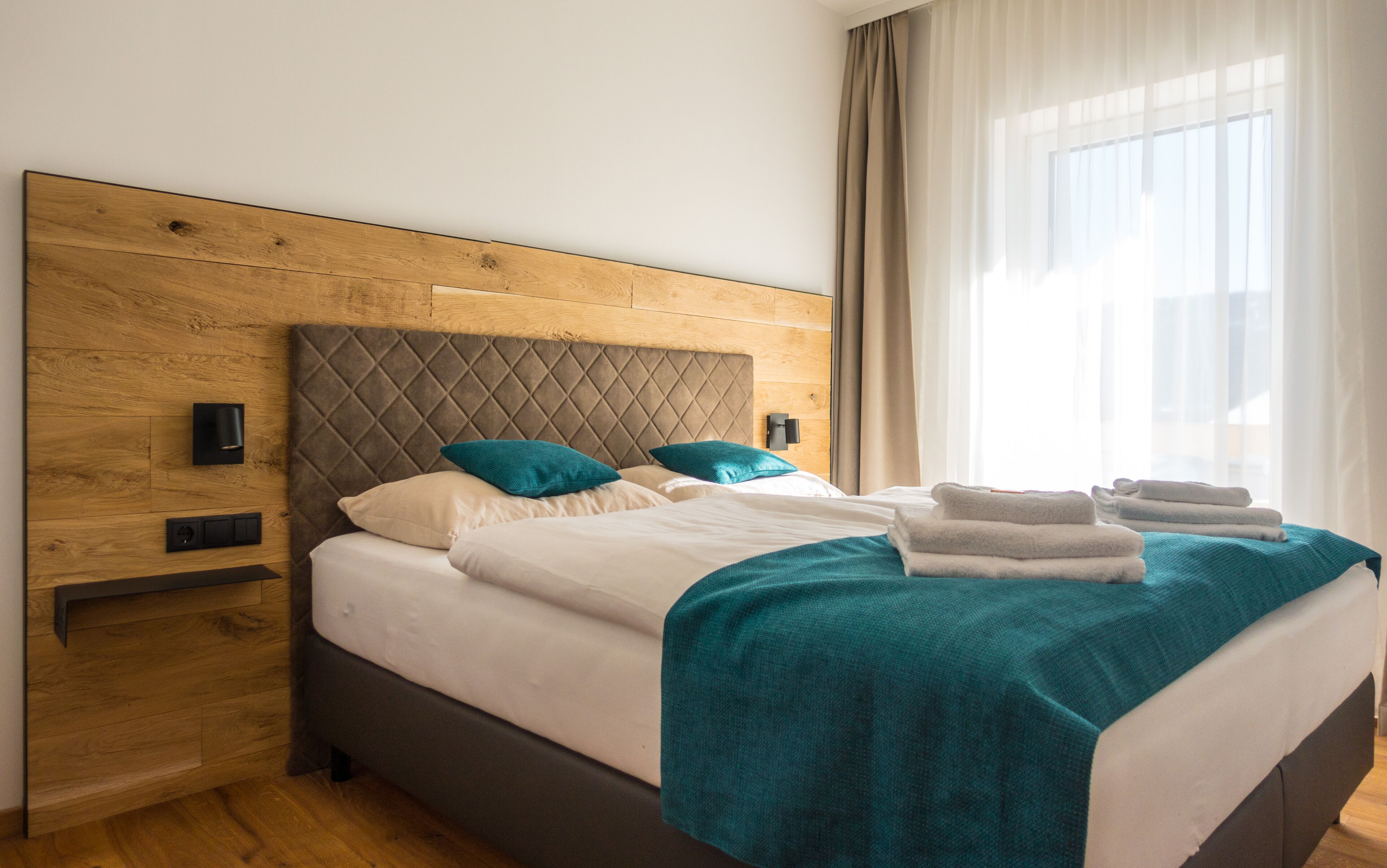 A neatly made bed with a wooden headboard, teal pillows, and a teal throw blanket.