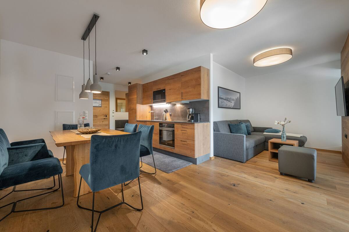 Una cocina moderna con armarios de madera, un comedor con sillas azules y un sofá gris en una sala de estar.