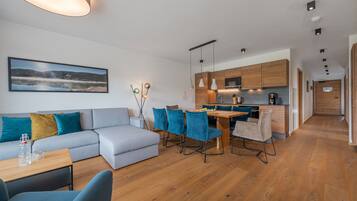 A modern living room with a sofa, a dining table with chairs, a kitchen area with wooden cabinets, and a hallway leading to another room.