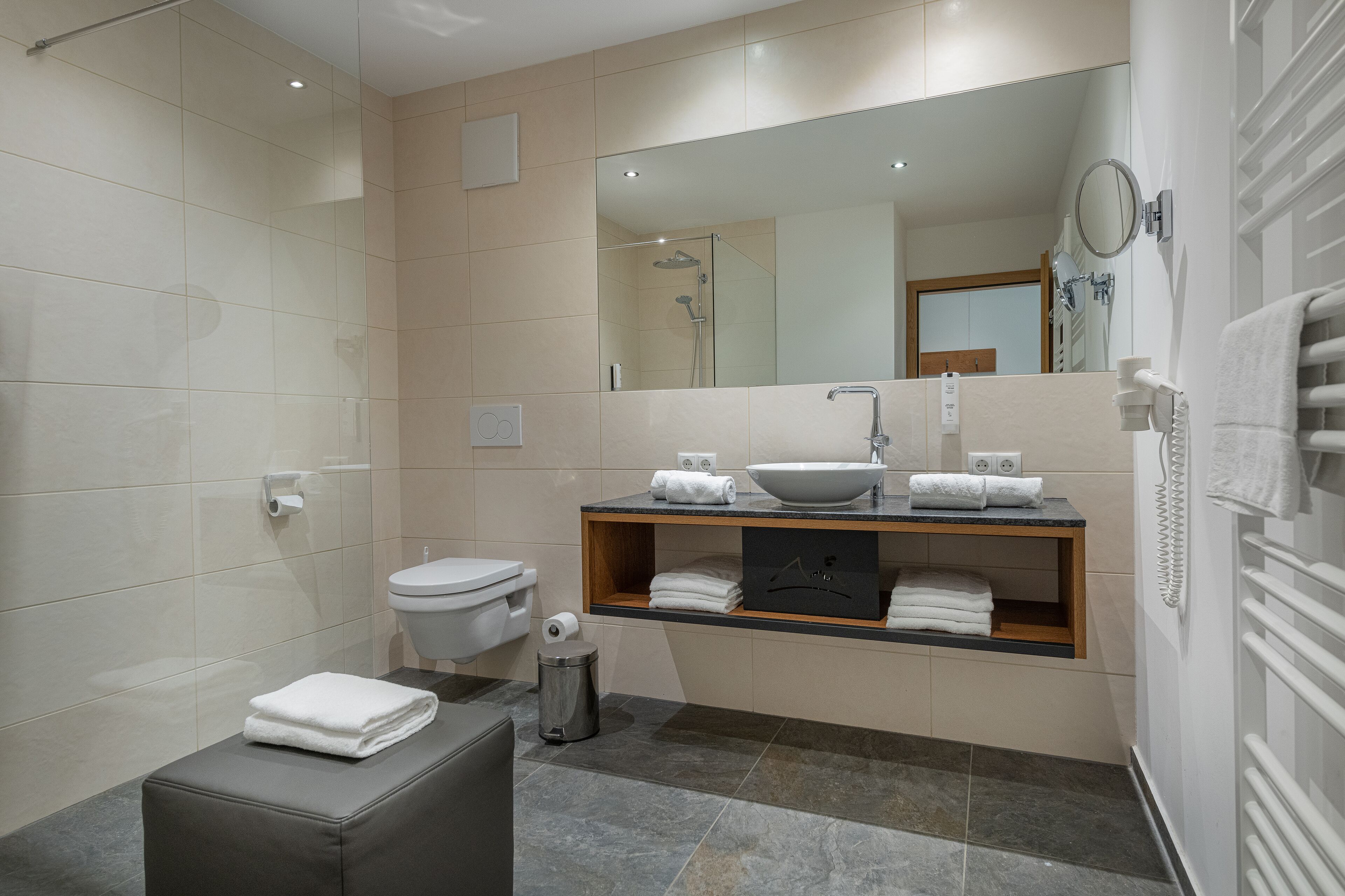 A modern bathroom with a toilet, sink, and shower. There is a towel bar with folded towels, a trash can, and a mirror above the sink.