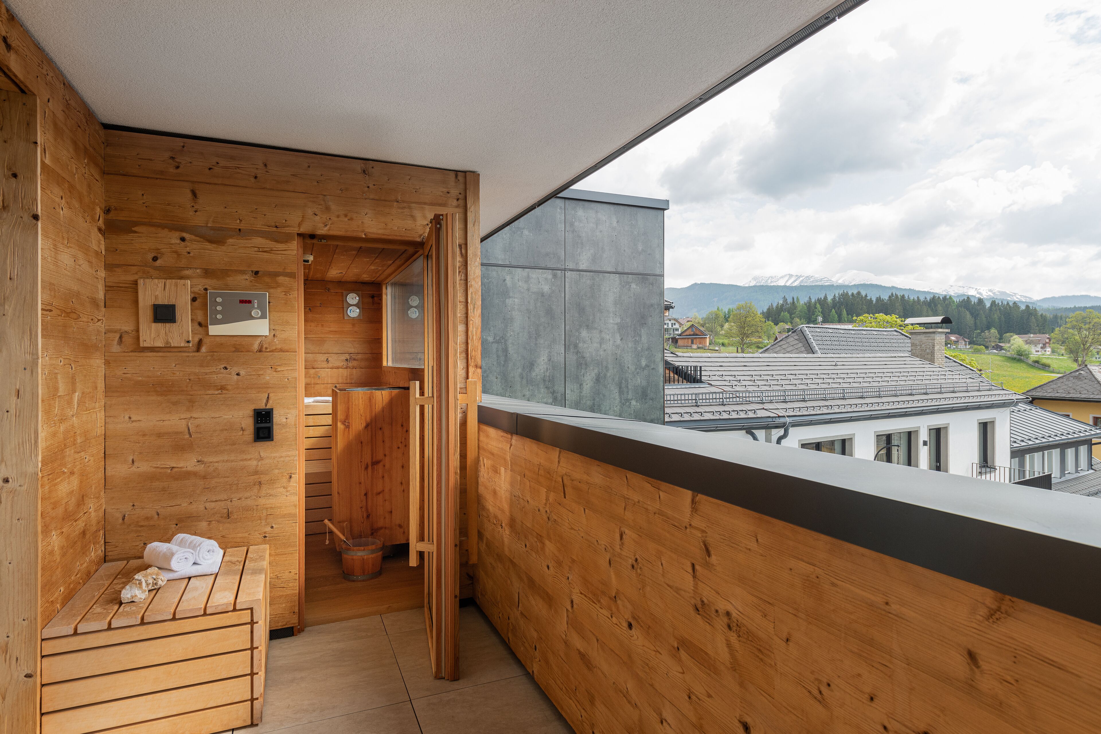 A wooden cabin with a sauna, a wooden bench, and a view of houses and mountains.