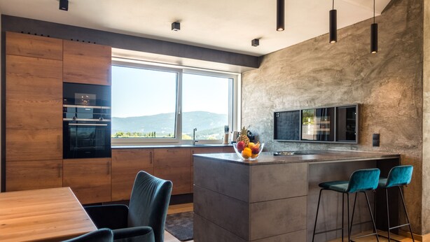 A modern kitchen with wooden cabinets, a large window, a central island with bar stools, and a fruit bowl. - AlpenParks Hotel & Apartment Carpe Solem (Mariapfarr)