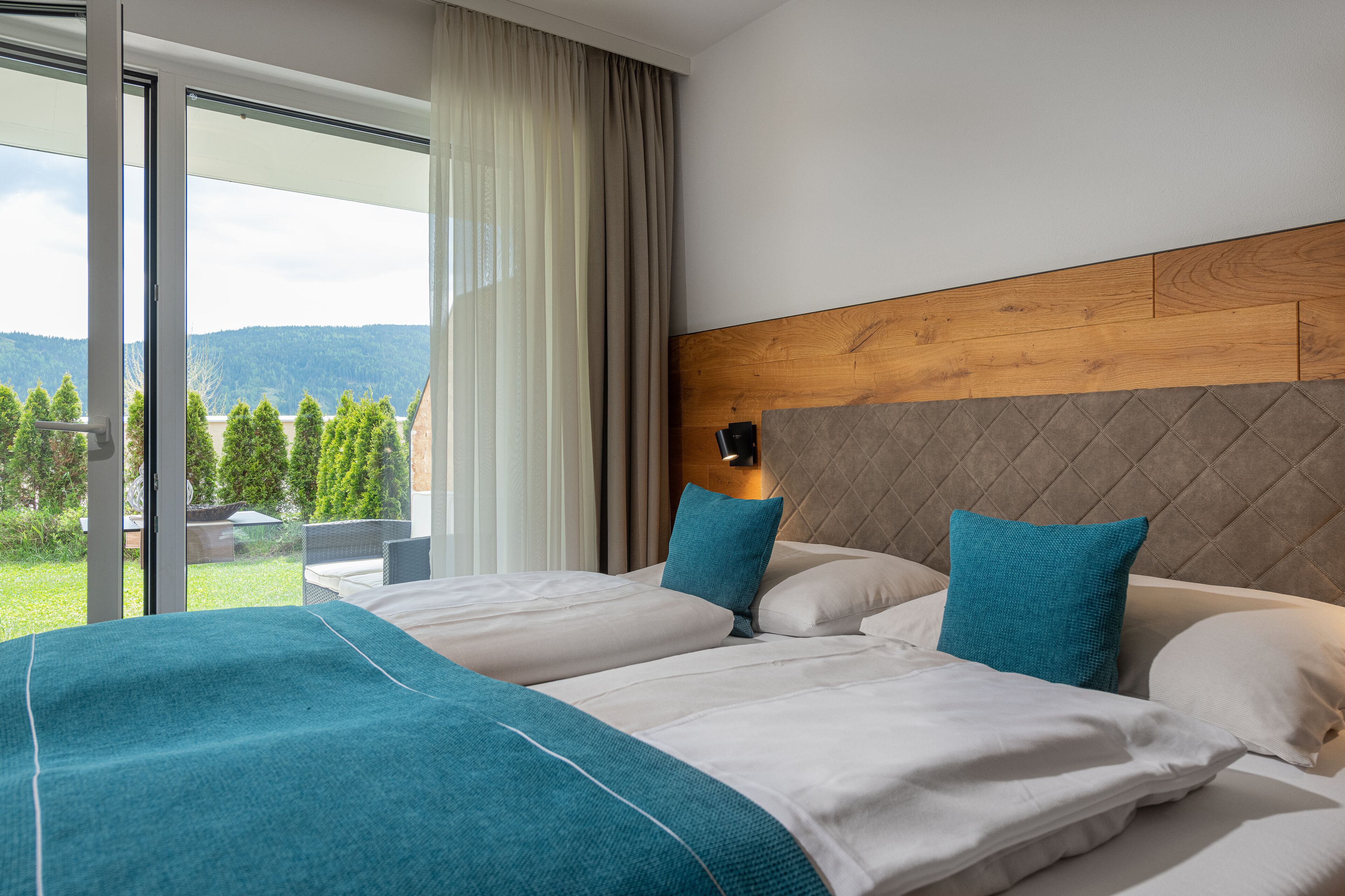 A hotel room with a large bed, a wooden headboard, and a view of greenery and mountains through the window.