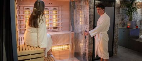 A modern spa area with a wooden bench and a glass enclosure, a man in a robe, and a woman in a robe sitting on the bench.